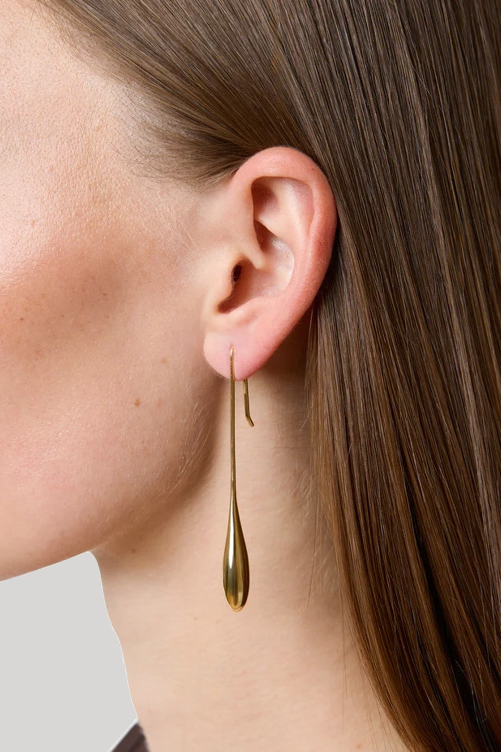 A close-up of a model's ear wearing a long, minimalist gold dangle earring with a teardrop-shaped drop and a small curved accent above it.