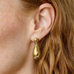 A close-up of a model's ear wearing a polished gold teardrop dangle earring with a small gold ball stud.