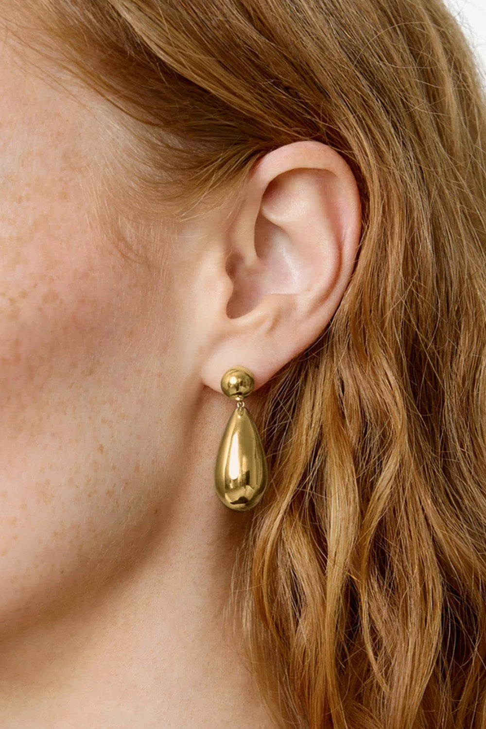 A close-up of a model's ear wearing a polished gold teardrop dangle earring with a small gold ball stud.