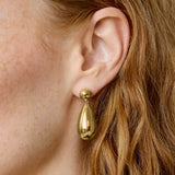 A close-up of a model's ear wearing a polished gold teardrop dangle earring with a small gold ball stud.