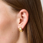 A close-up side profile of a woman's ear wearing a small gold huggie hoop earring with a teardrop-shape.