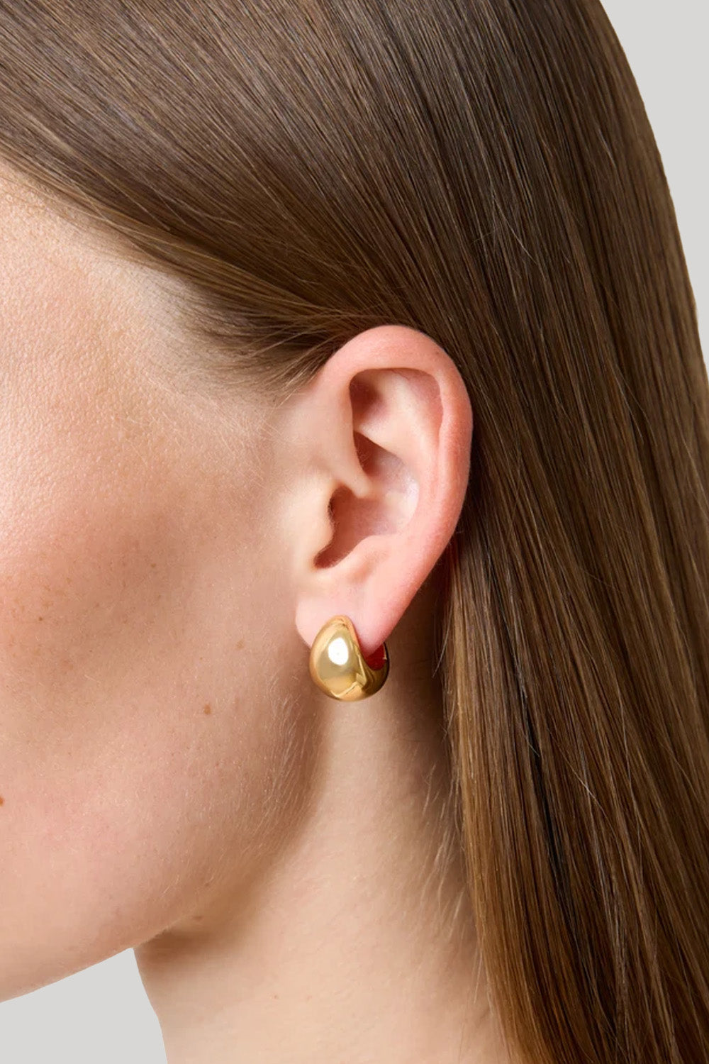 A close-up side profile of a woman's ear wearing a small gold huggie hoop earring with a teardrop-shape.