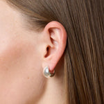 A close-up side profile of a model's ear wearing a small silver hoop earring with a teardrop-shaped pearl-like drop, against brown hair and a light gray background.