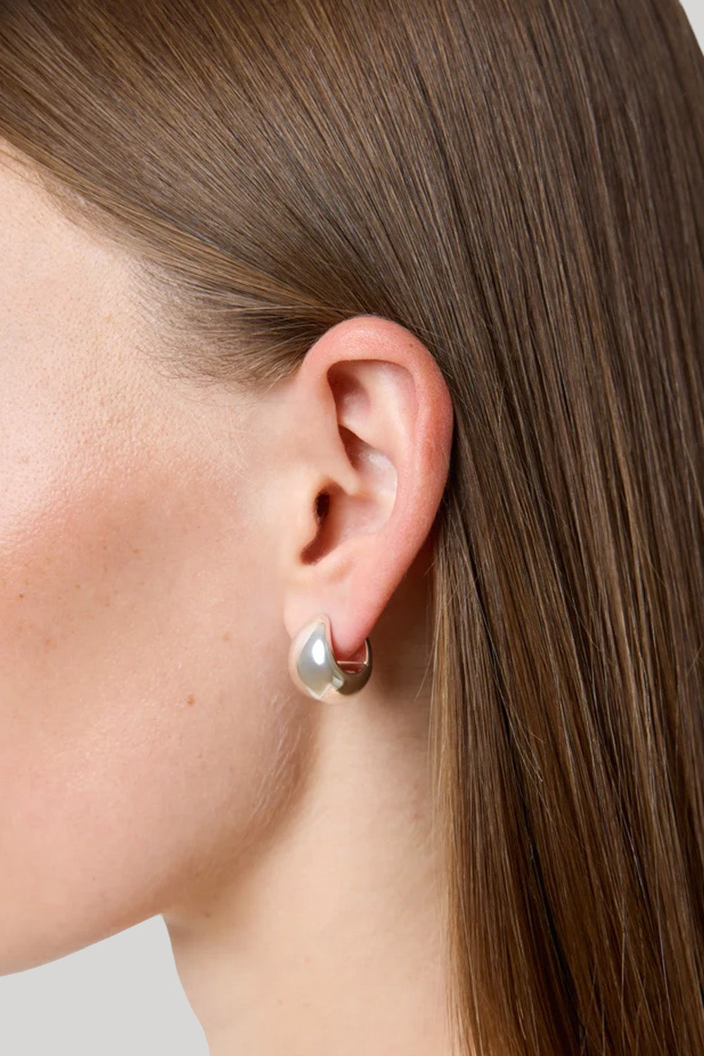 A close-up side profile of a model's ear wearing a small silver hoop earring with a teardrop-shaped pearl-like drop, against brown hair and a light gray background.