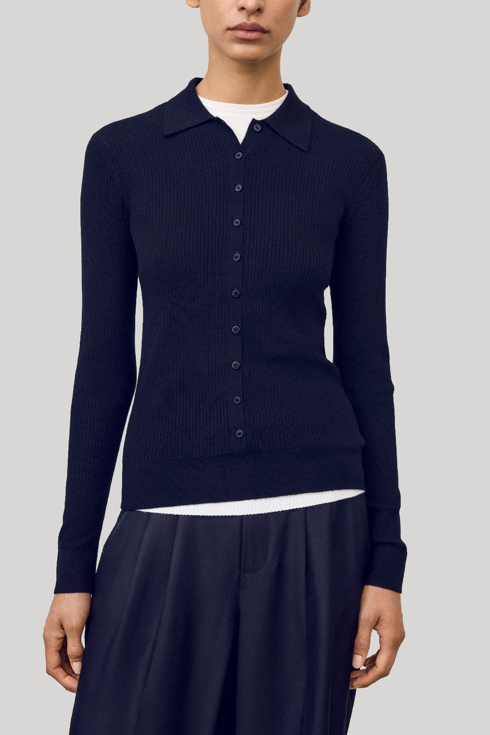 A model wearing a navy ribbed knit polo cardigan over a white undershirt and dark wide-leg trousers, photographed against a solid light gray studio background.