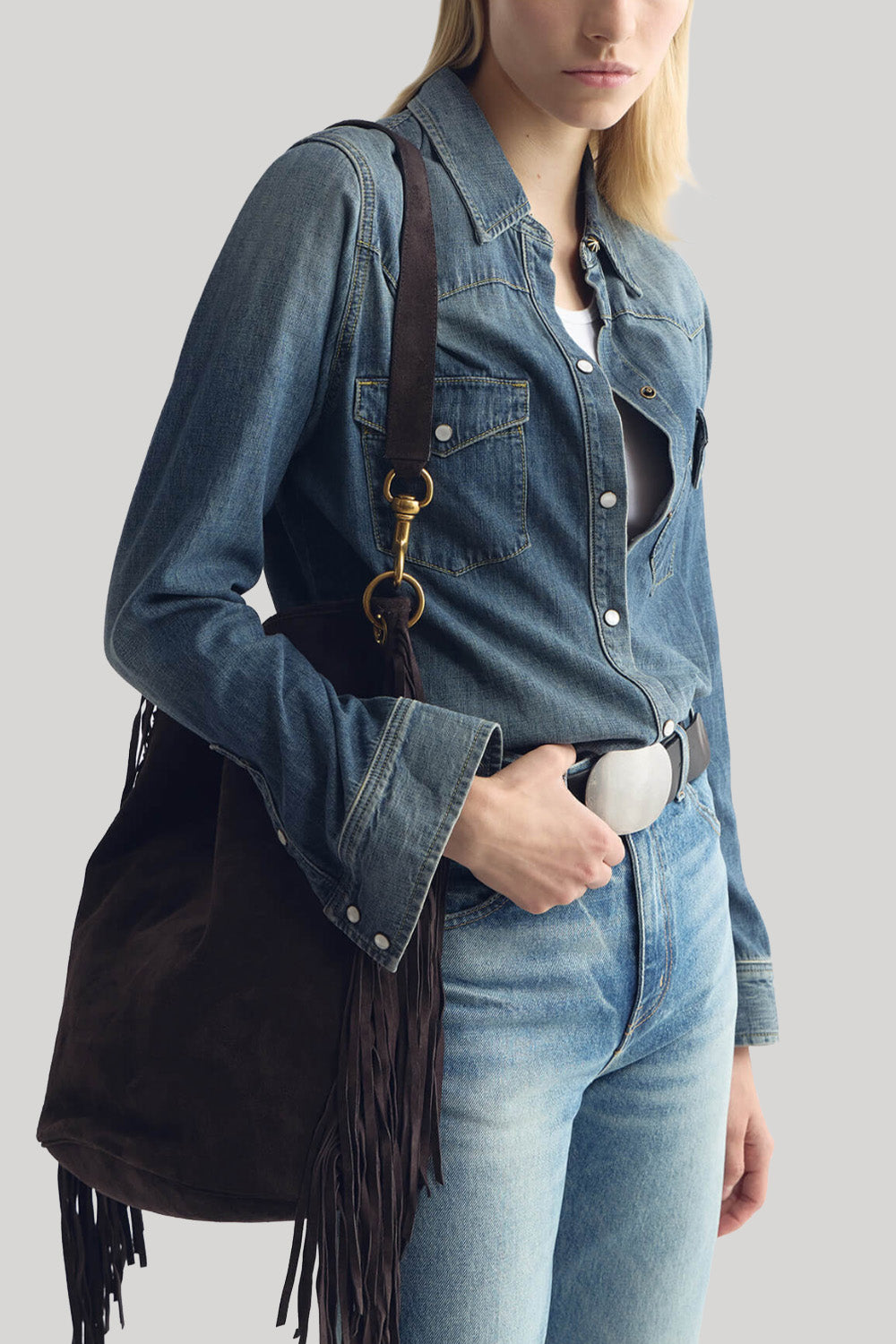 A model wearing a blue denim shirt and jeans, carrying a dark brown suede fringe shoulder bag with gold-tone hardware from Nili Lotan against a light gray background.