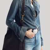 A model wearing a blue denim shirt and jeans, carrying a dark brown suede fringe shoulder bag with gold-tone hardware from Nili Lotan against a light gray background.