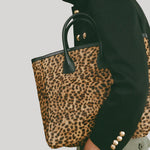 A moel wearing a black jacket and olive pants carries a leopard-print tote bag with black handles and gold-tone hardware from Nili Lotan against a light gray background.