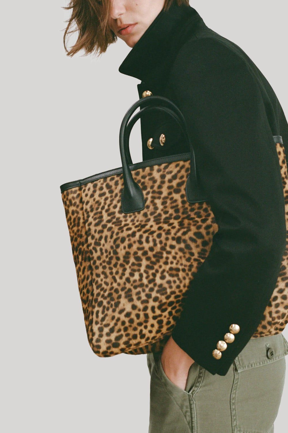 A moel wearing a black jacket and olive pants carries a leopard-print tote bag with black handles and gold-tone hardware from Nili Lotan against a light gray background.