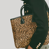 A moel wearing a black jacket and olive pants carries a leopard-print tote bag with black handles and gold-tone hardware from Nili Lotan against a light gray background.
