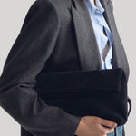 A model wearing a dark gray plaid blazer over a light blue button-down shirt, holding a navy velvet clutch bag from Nili Lotan against their torso on a plain grey background