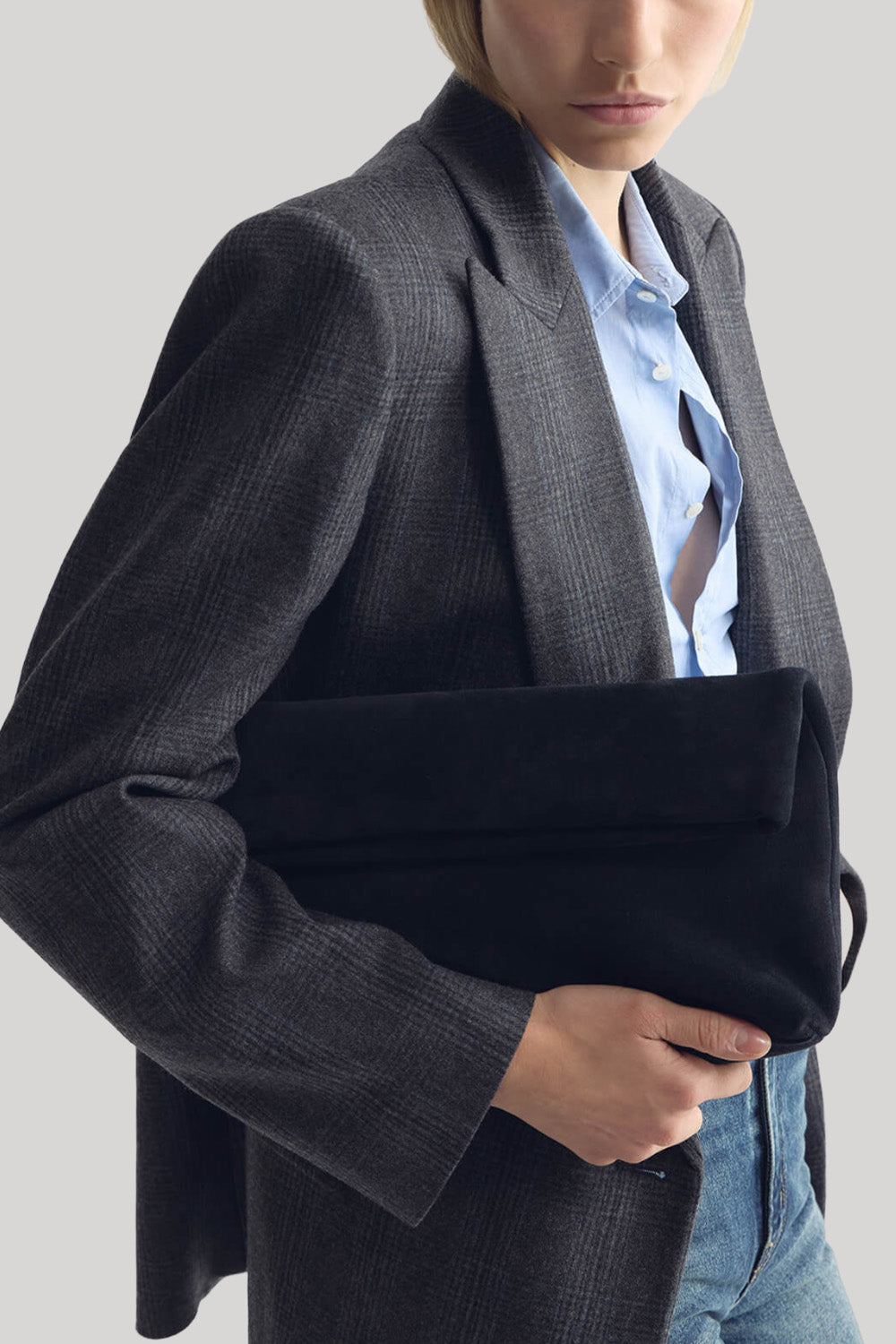 A model wearing a dark gray plaid blazer over a light blue button-down shirt, holding a navy velvet clutch bag from Nili Lotan against their torso on a plain grey background
