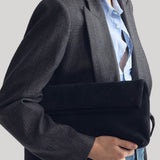 A model wearing a dark gray plaid blazer over a light blue button-down shirt, holding a navy velvet clutch bag from Nili Lotan against their torso on a plain grey background