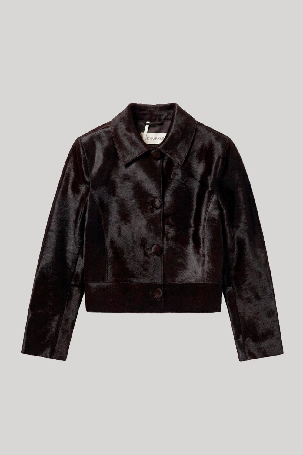 A dark brown cropped pony hair leathre jacket with a pointed collar and three-quarter sleeves, shown flat on a light gray background. The jacket is from Nour Hammour