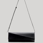 A black leather shoulder bag with an asymmetrical diagonal flap and a thin black strap, shown against a solid light gray background.