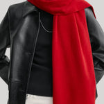 A close-up of a model wearing a vibrant red knit scarf draped over a black leather jacket, black top, and white pants against a light gray background.
