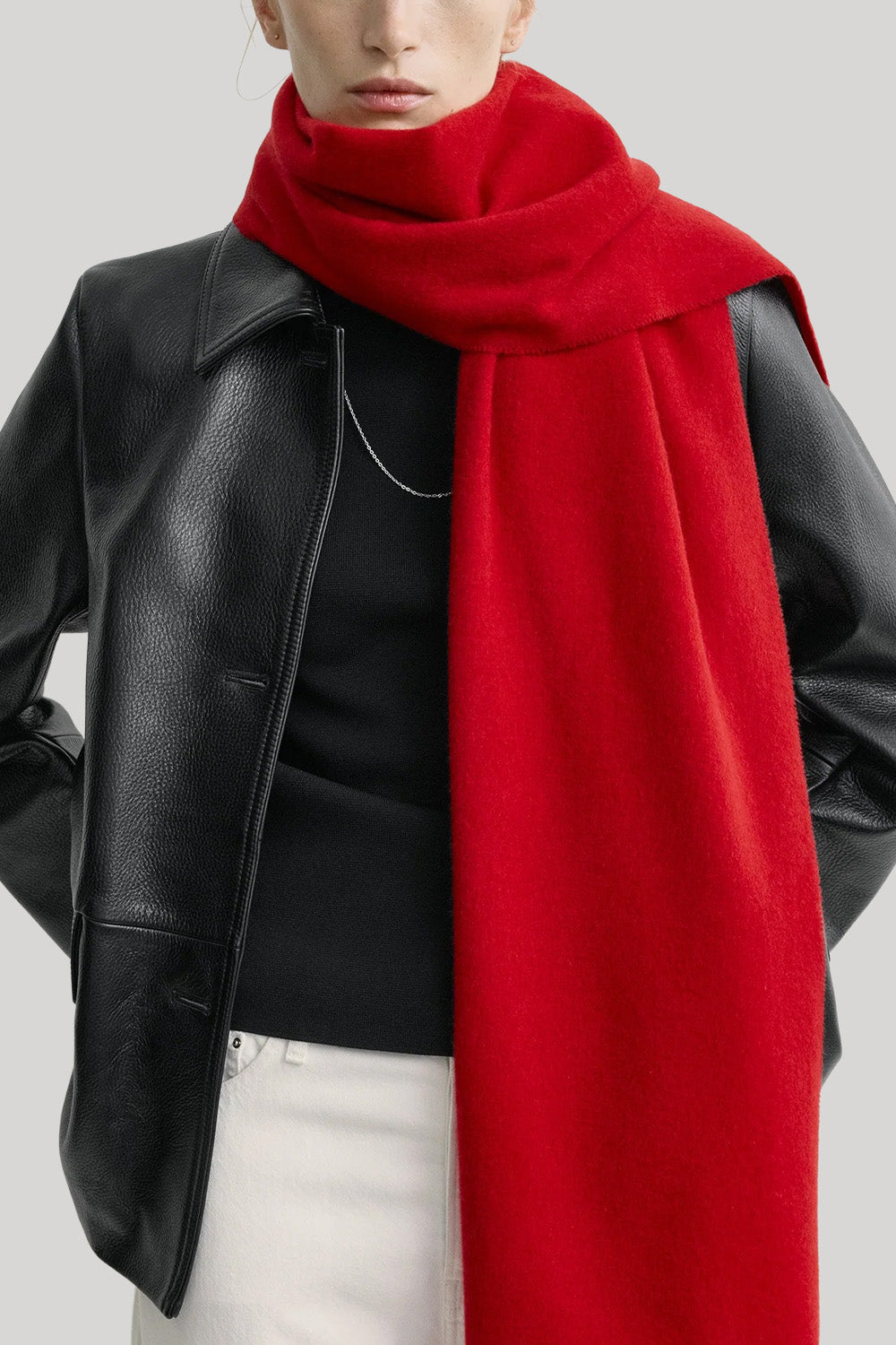 A close-up of a model wearing a vibrant red knit scarf draped over a black leather jacket, black top, and white pants against a light gray background.