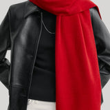 A close-up of a model wearing a vibrant red knit scarf draped over a black leather jacket, black top, and white pants against a light gray background.