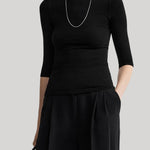 A model wearing a black three-quarter sleeve top and matching black wide-leg trousers, accessorized with a silver chain necklace and holding a black handbag, set against a solid light gray background.