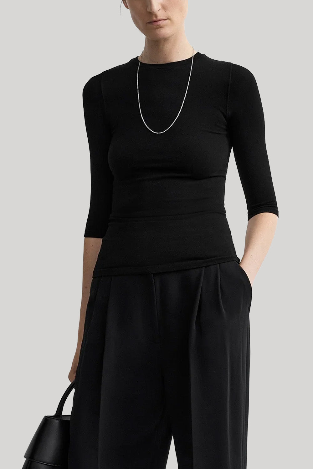 A model wearing a black three-quarter sleeve top and matching black wide-leg trousers, accessorized with a silver chain necklace and holding a black handbag, set against a solid light gray background.