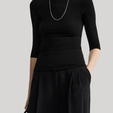 A model wearing a black three-quarter sleeve top and matching black wide-leg trousers, accessorized with a silver chain necklace and holding a black handbag, set against a solid light gray background.