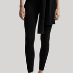 A model wearing black leggings with a matching black knit wrap tied at the waist, paired with black ballet flats, against a solid light gray background.