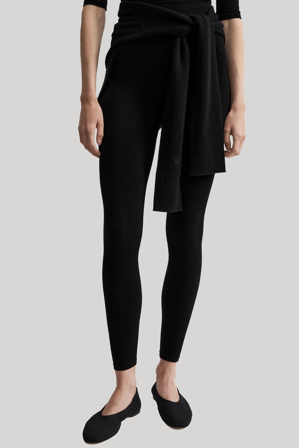 A model wearing black leggings with a matching black knit wrap tied at the waist, paired with black ballet flats, against a solid light gray background.