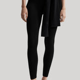 A model wearing black leggings with a matching black knit wrap tied at the waist, paired with black ballet flats, against a solid light gray background.