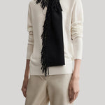 A model wearing a cream-colored knit sweater and matching trousers, styled with a black fringed scarf, against a solid light gray background.