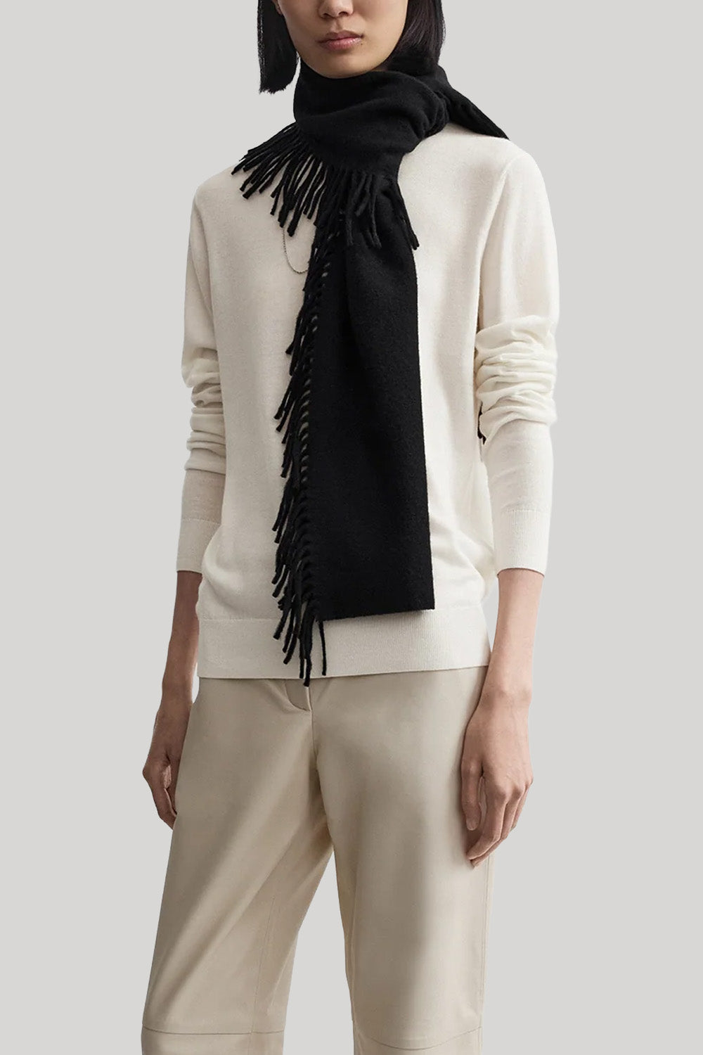 A model wearing a cream-colored knit sweater and matching trousers, styled with a black fringed scarf, against a solid light gray background.
