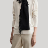 A model wearing a cream-colored knit sweater and matching trousers, styled with a black fringed scarf, against a solid light gray background.
