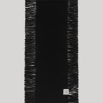 A black rectangular scarf with fringed edges, laid flat against a light gray background. A small square white brand tag is visible near the bottom right corner.