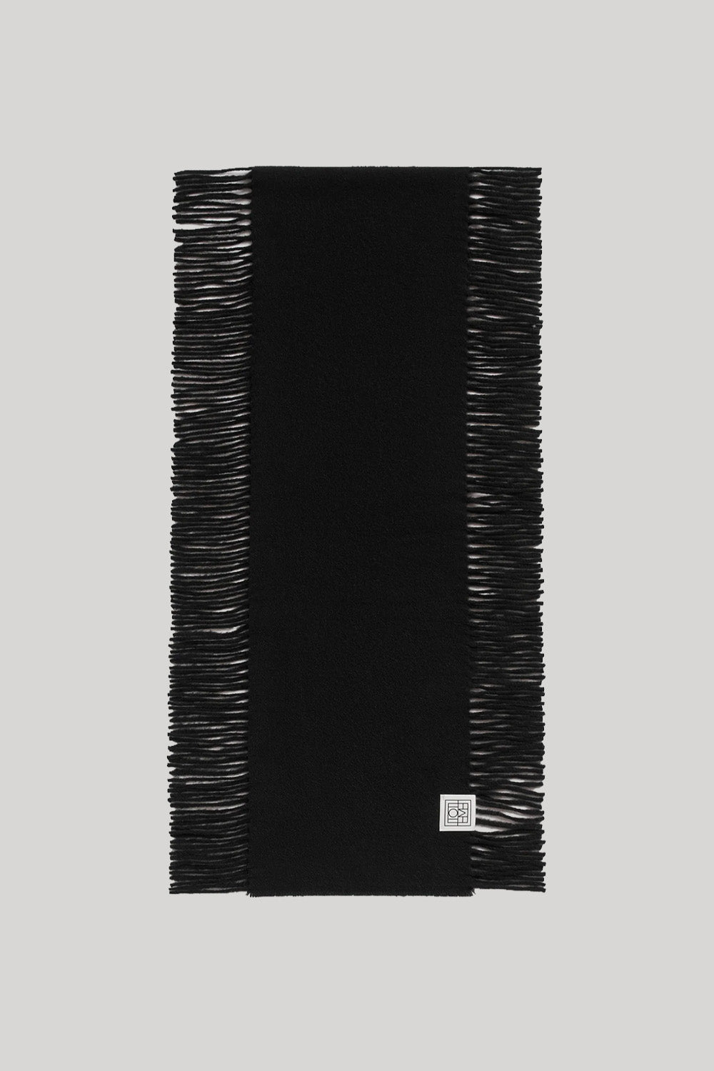 A black rectangular scarf with fringed edges, laid flat against a light gray background. A small square white brand tag is visible near the bottom right corner.