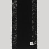 A black rectangular scarf with fringed edges, laid flat against a light gray background. A small square white brand tag is visible near the bottom right corner.