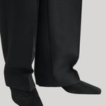 Close-up of black wide-leg trousers worn with black pointed-toe shoes against a light gray background.
