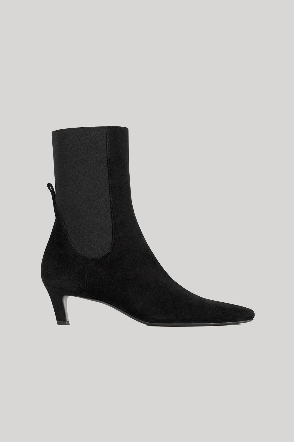 A black suede ankle boot with a square toe and a slim black kitten heel, featuring a black elastic side panel, shown against a solid light gray background.