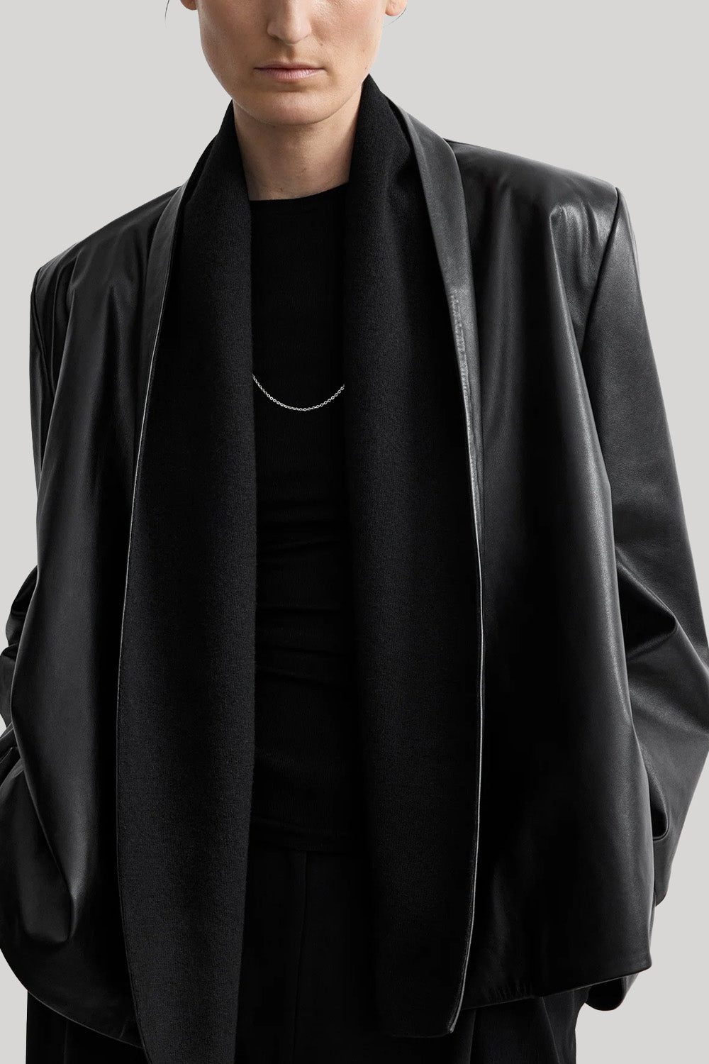 A close-up of a model wearing a black leather-look shawl-collar jacket over a black top and black scarf, with a thin silver necklace visible against the collar.