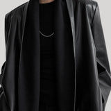 A close-up of a model wearing a black leather-look shawl-collar jacket over a black top and black scarf, with a thin silver necklace visible against the collar.