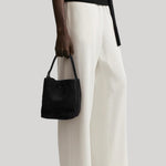 A model wearing off-white wide-leg trousers, a black top, and black shoes, holding a black suede bucket bag  from Toteme against a light gray background. 