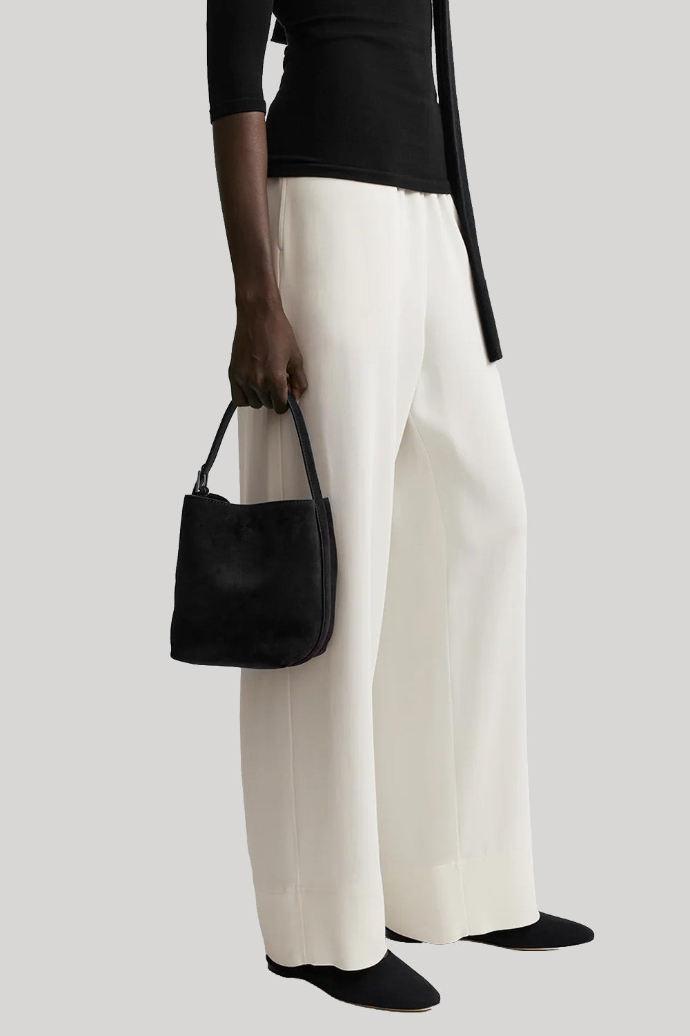 A model wearing off-white wide-leg trousers, a black top, and black shoes, holding a black suede bucket bag  from Toteme against a light gray background. 