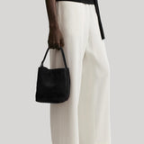 A model wearing off-white wide-leg trousers, a black top, and black shoes, holding a black suede bucket bag  from Toteme against a light gray background. 