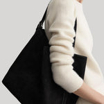 A person wearing a cream knit sweater and light pants carries a large black shoulder bag with a wide slouchy silhouette and a wide strap against a light gray studio background.