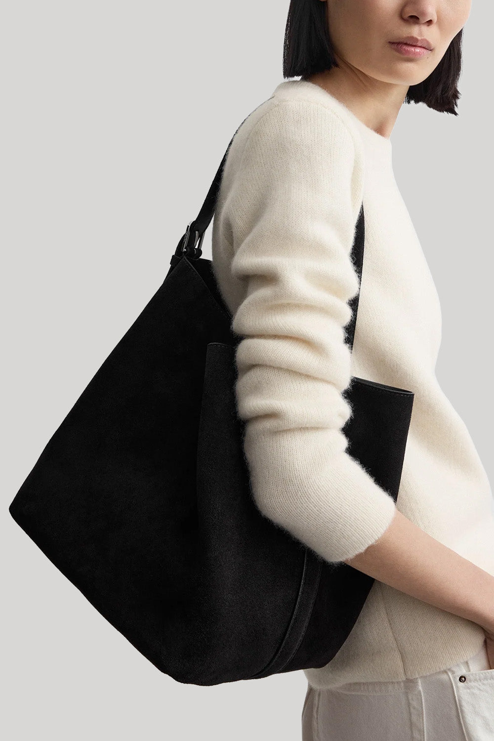 A person wearing a cream knit sweater and light pants carries a large black shoulder bag with a wide slouchy silhouette and a wide strap against a light gray studio background.