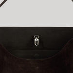 Close-up of a dark brown suede shoulder bag from Toteme with a silver metal D-ring clasp on a light gray background.