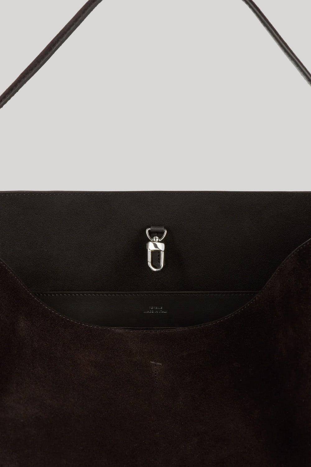 Close-up of a dark brown suede shoulder bag from Toteme with a silver metal D-ring clasp on a light gray background.