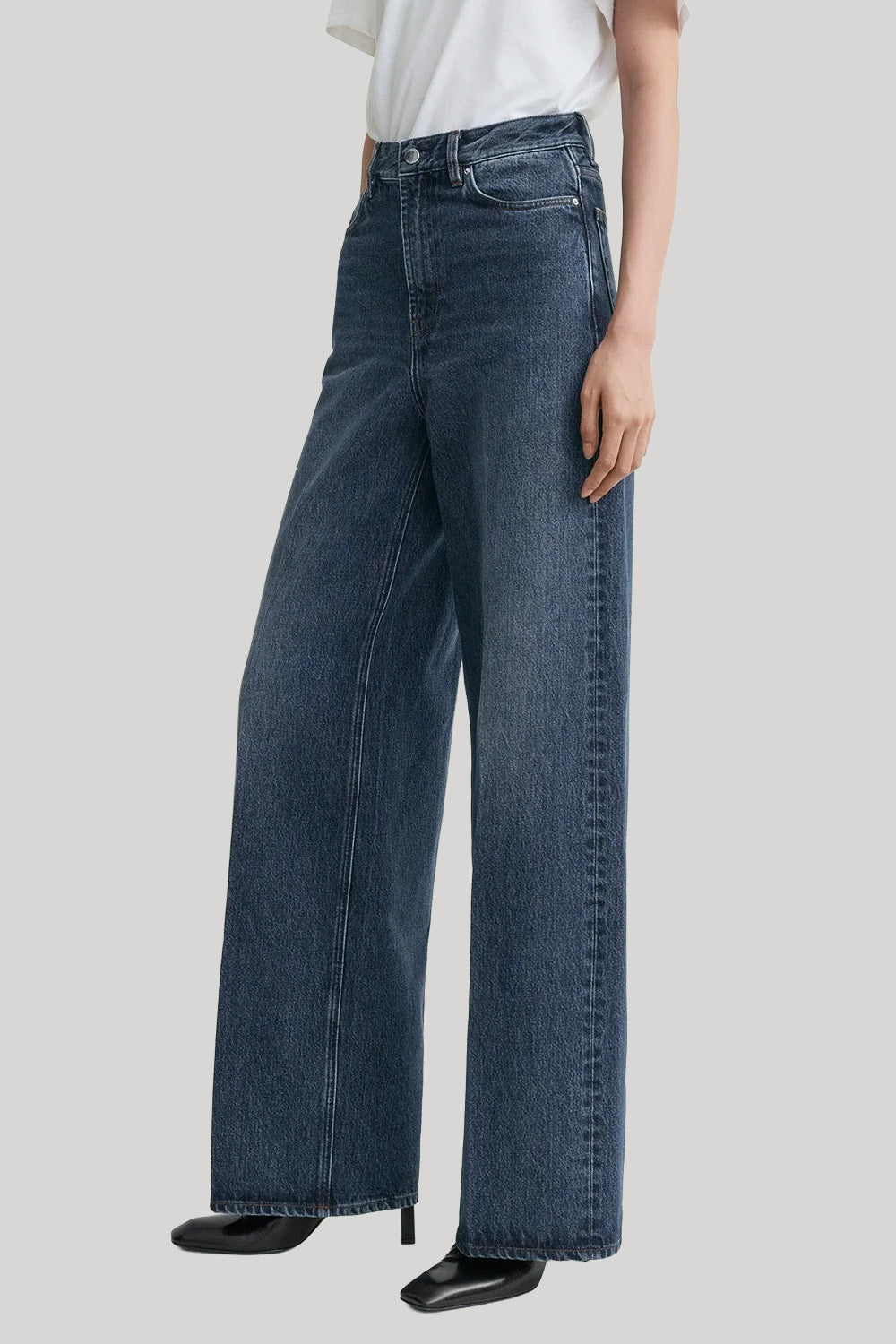 A model wearing a pair of high-waisted, wide-leg blue denim jeans from Toteme, styled with a white top and black pointed-toe heels against a light gray studio background.