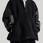 A model wearing a black oversized button-front varsity jacket with contrasting black leather sleeves, styled over a black turtleneck, white collared shirt, and black trousers, holding a black textured clutch against a solid gray background.