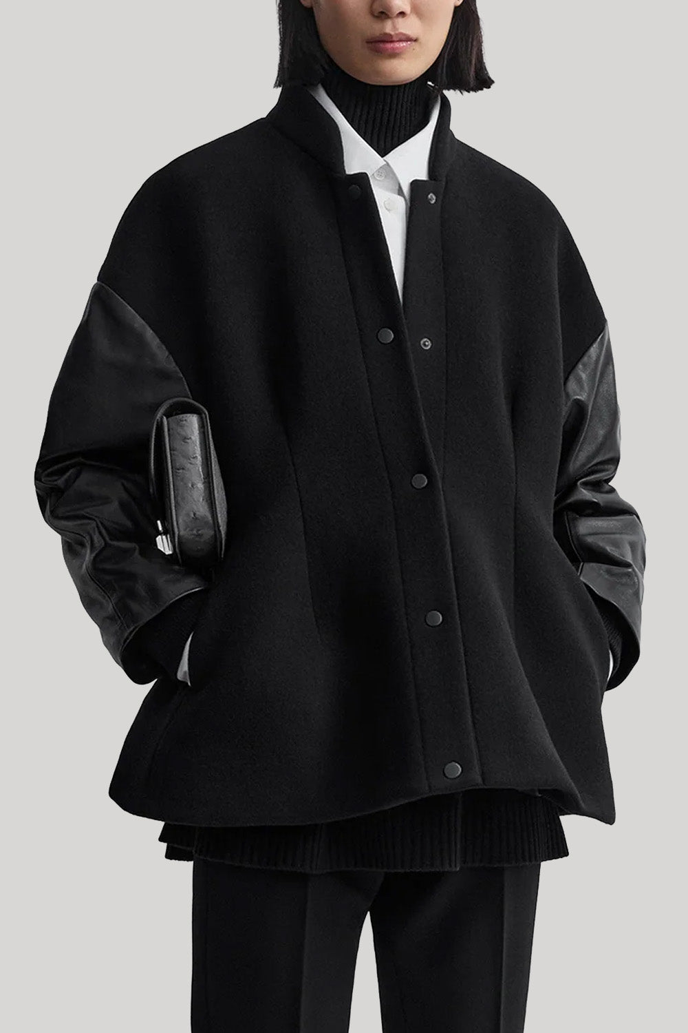 A model wearing a black oversized button-front varsity jacket with contrasting black leather sleeves, styled over a black turtleneck, white collared shirt, and black trousers, holding a black textured clutch against a solid gray background.