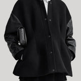A model wearing a black oversized button-front varsity jacket with contrasting black leather sleeves, styled over a black turtleneck, white collared shirt, and black trousers, holding a black textured clutch against a solid gray background.
